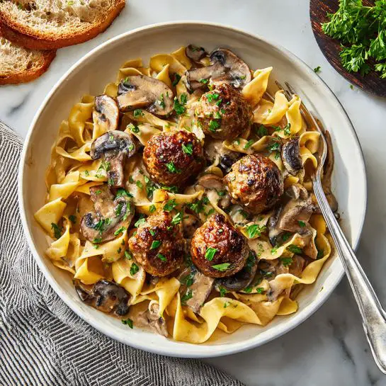 The dish shows a white bowl filled with wide, soft egg noodles that are light golden-yellow in color, mixed with thick slices of brown, sautéed mushrooms scattered evenly. On top sit several browned meatballs with a slightly rough texture, covered in a creamy, beige sauce all over the noodles and mushrooms. Small, bright green parsley leaves are sprinkled on top for color. A silver fork rests inside the bowl on the right side. The bowl is on a white marbled table with a striped cloth partially visible on the bottom left corner and a dark bread slice and parsley sprig on the top right edges. Photo taken with an iphone --ar 4:5 --v 7