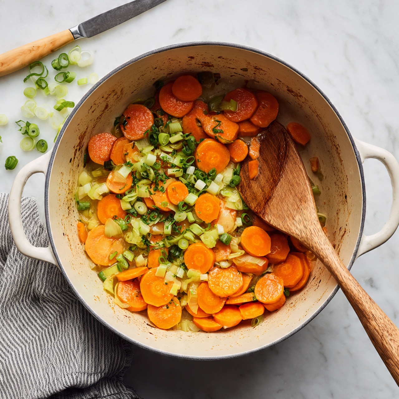 Inside a deep white pot, there is a mix of sliced orange carrots and chopped light green scallions. The vegetables sit mostly on one side of the pot, showing some browning bits on the beige interior of the pot. A wooden spatula lies inside the pot on the right side, touching the vegetables. Around the pot, on a white marbled surface, there are a few scattered pieces of sliced green onions and a knife with a light wood handle near the top left. A gray and white striped cloth peeks out from the bottom right corner. photo taken with an iphone --ar 4:5 --v 7