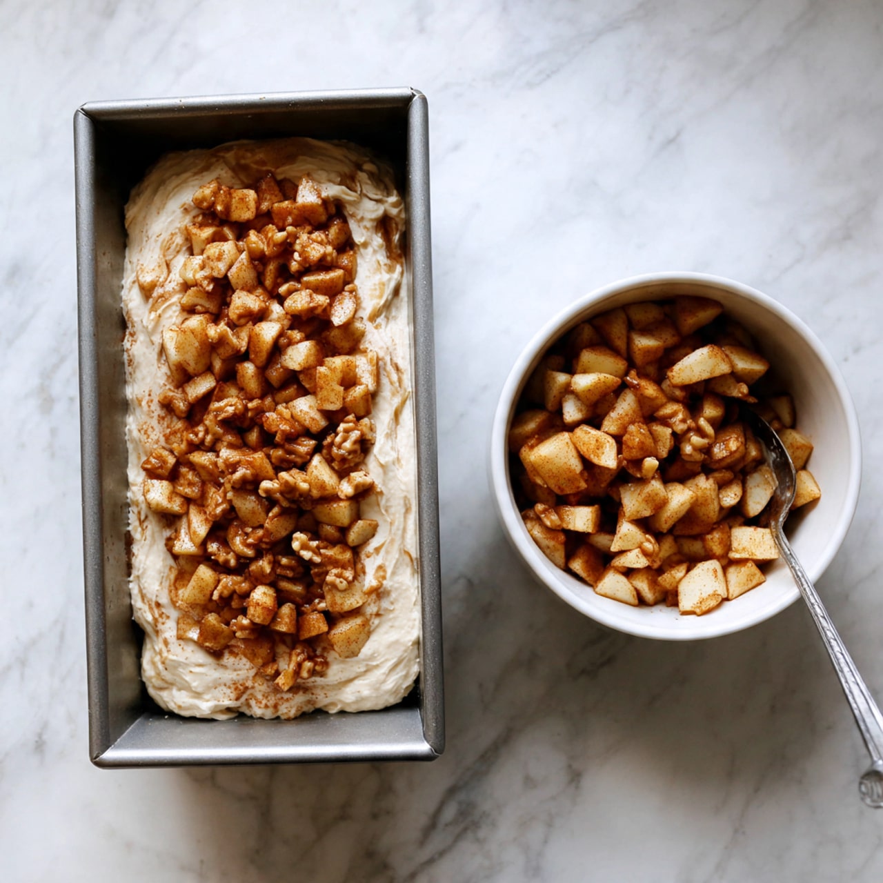 A metal rectangular loaf pan holds a thick, pale cream batter spread evenly across the bottom. On top of this batter, there is a layer of small, diced apple pieces coated in a cinnamon spice, giving them a warm brown color. To the right of the pan, a white bowl contains more apple pieces and a silver spoon resting inside, all placed on a white marbled surface. Photo taken with an iphone --ar 4:5 --v 7