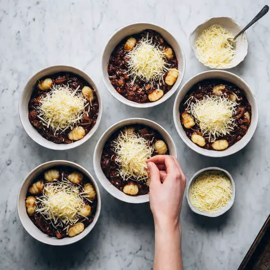 The image shows six white round bowls arranged on a dark baking tray placed on a white marbled surface. Each bowl contains a dark brown mixture with chunks of gnocchi and onion visible at the bottom. On top of this mixture, there is a layer of shredded light yellow cheese being sprinkled. A woman's hand is seen adding cheese to one of the bowls at the bottom left. Nearby, on the white marbled surface, there are two small white bowls, one filled with shredded cheese and the other with grated cheese. Photo taken with an iphone --ar 4:5 --v 7