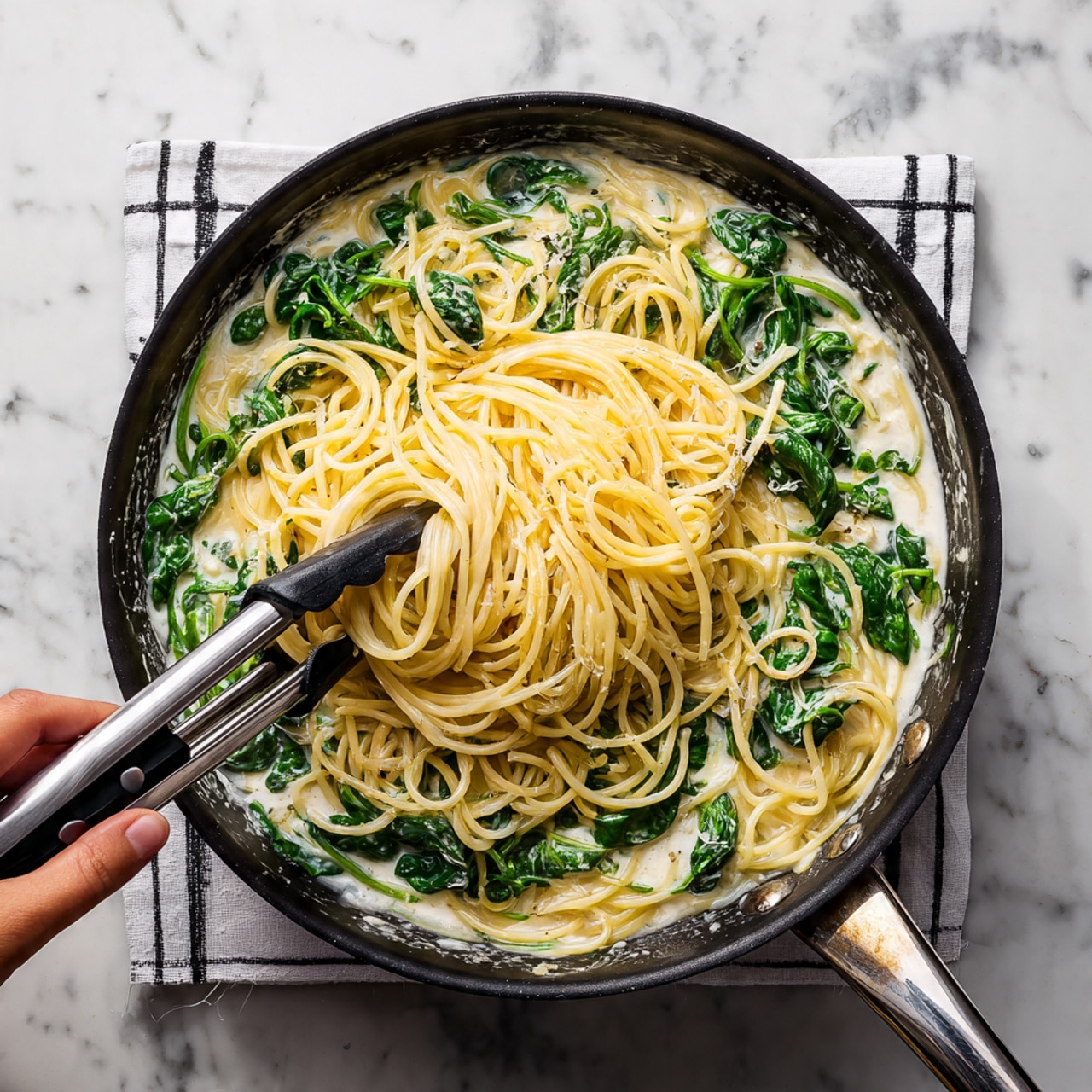 A black pan filled with creamy white sauce at the bottom holds a layer of fresh green spinach leaves all around. On top, a woman’s hand uses silver tongs with black tips to lift a nest of pale yellow spaghetti noodles that are mixed with some of the spinach, showing steam rising from the hot pasta. The pan sits on a white cloth with a black grid pattern on a white marbled surface. Photo taken with an iphone --ar 4:5 --v 7