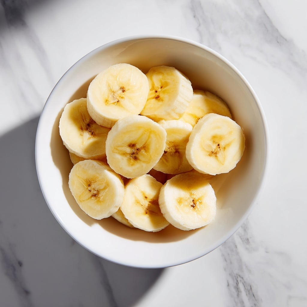Two peeled bananas are placed side by side on a simple white round plate, their soft pale yellow flesh showing natural brown specks along the middle lines. The plate rests on a white marbled surface, and a light white cloth is partly visible at the bottom right corner. The image is bright and clean with minimal shadows, giving a fresh and natural feel. photo taken with an iphone --ar 4:5 --v 7