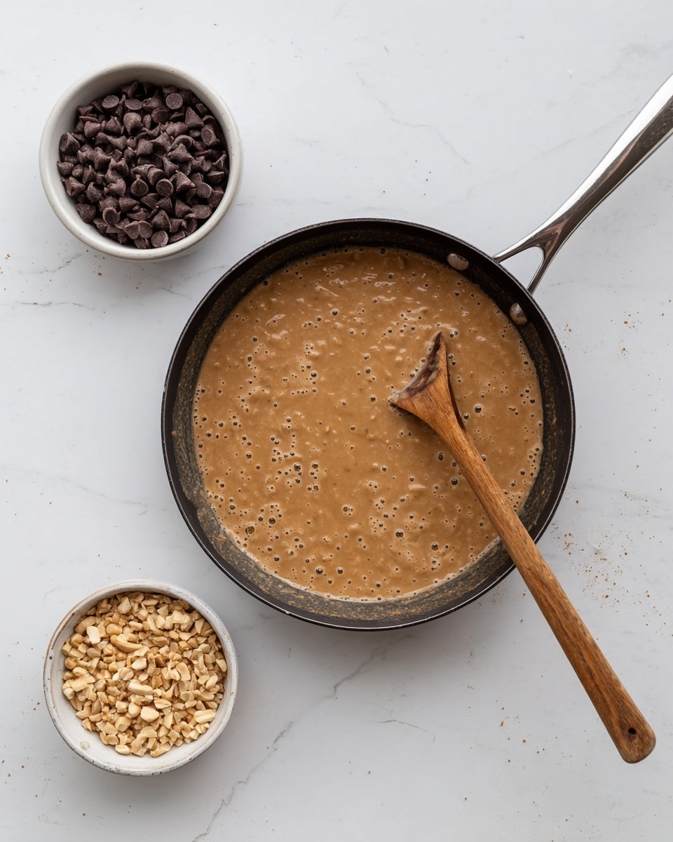 A black frying pan with a silver handle holds a thick, light brown mixture with a bumpy texture and small bubbles on top. A wooden spoon with a smooth surface rests inside the pan, partially submerged in the mixture. To the left of the pan, there is a small white bowl filled with dark brown chocolate chips, and to the right, another small white bowl contains chopped light brown nuts. All items are set on a white marbled surface. Photo taken with an iphone --ar 4:5 --v 7