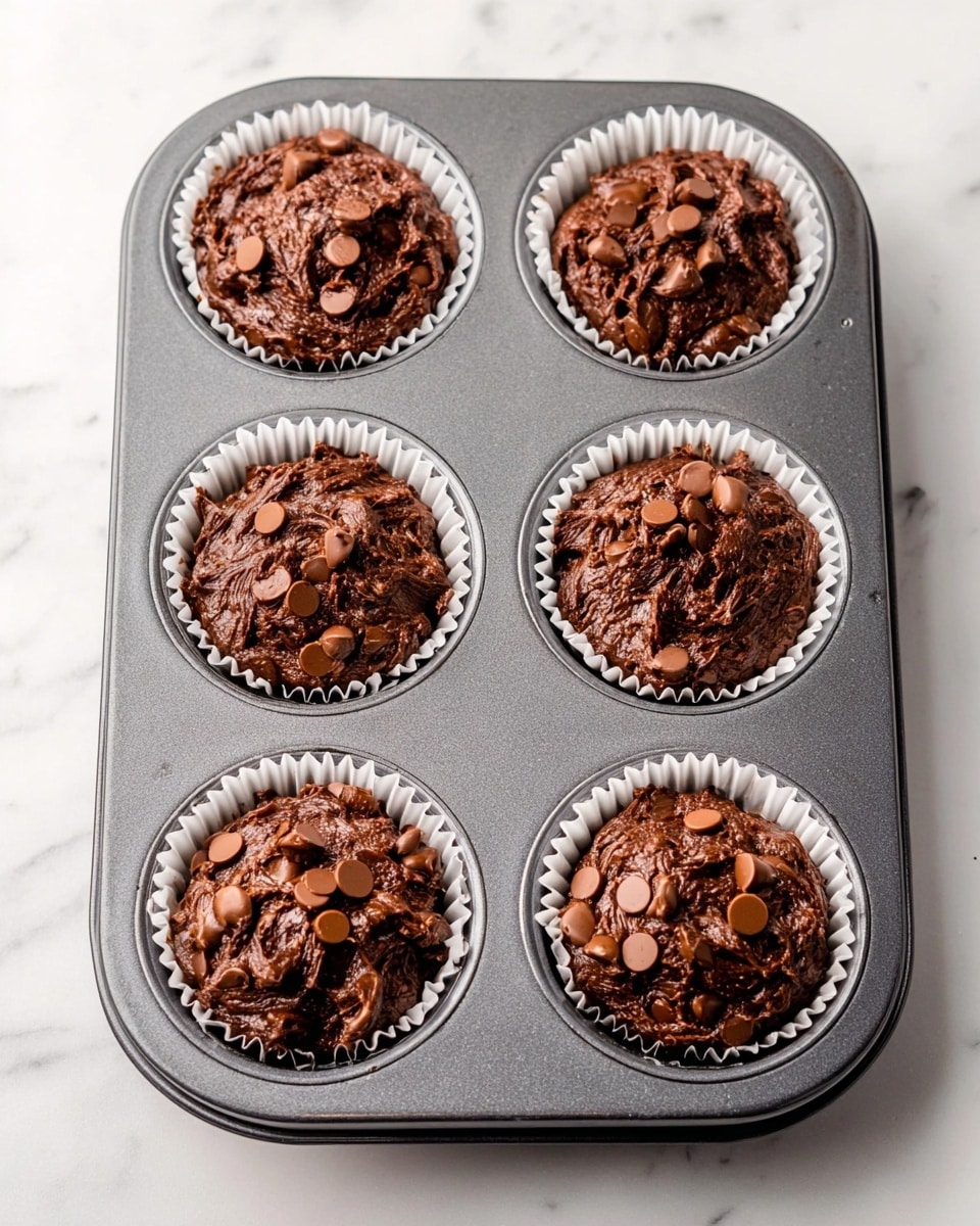 A dark gray metal muffin tray holds six white paper liners filled with thick, rich chocolate batter. Each liner is filled almost to the top with the dense, glossy batter that has a slightly rough texture, showing swirls and uneven peaks. Scattered on each mound of batter are flat, light brown chocolate chips that add a bit of texture contrast. The muffin tray rests on a clean white marbled surface. photo taken with an iphone --ar 4:5 --v 7