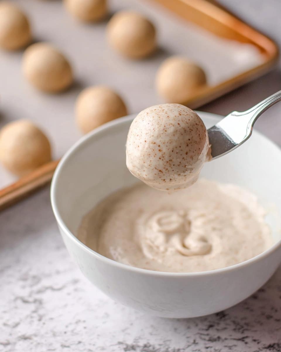 A fork holds a smooth, round ball covered in a creamy, light beige mixture with tiny brown specks. This ball is above a white bowl filled with the same creamy mixture, which appears thick and slightly textured. In the background, multiple similar uncooked beige balls sit spaced out on a baking sheet. The scene is set on a white marbled textured surface. photo taken with an iphone --ar 4:5 --v 7