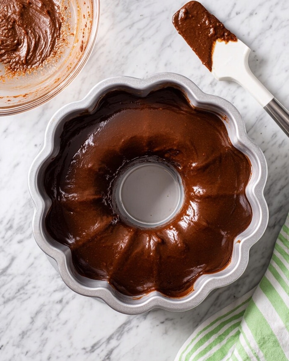 A clear glass bowl with some brown chocolate batter residue inside is placed on a white marbled surface. Next to it is a small white spatula with brown chocolate batter coating the blade. In the center of the image is a silver bundt cake pan filled with smooth, dark brown chocolate batter, evenly spread and shiny. The bundt pan has a fluted edge and a hollow center. A corner of a green and white striped cloth is slightly visible near the bottom right side of the image. photo taken with an iphone --ar 4:5 --v 7