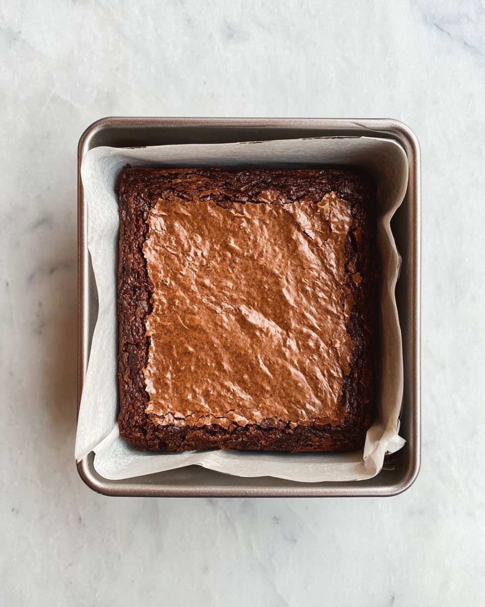 A square baked brownie with a cracked, textured top layer of rich brown color sits inside a metal baking pan lined with white parchment paper. The brownie fills the pan evenly, showing slightly darker edges and a soft middle. The pan rests on a white marbled surface. photo taken with an iphone --ar 4:5 --v 7