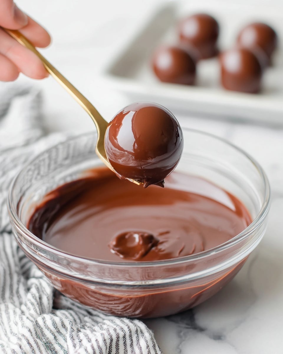 A shiny round chocolate ball is being held above a glass bowl filled with smooth melted milk chocolate that has a glossy texture. The chocolate ball is coated evenly in the melted chocolate, giving it a thick, rich dark brown layer. The bowl sits on a white marbled surface with a striped cloth partially visible underneath. In the background, there is a white tray holding three more uncoated chocolate balls, softly blurred to keep focus on the foreground. A woman's hand is gently holding a gold fork that supports the chocolate ball. photo taken with an iphone --ar 4:5 --v 7