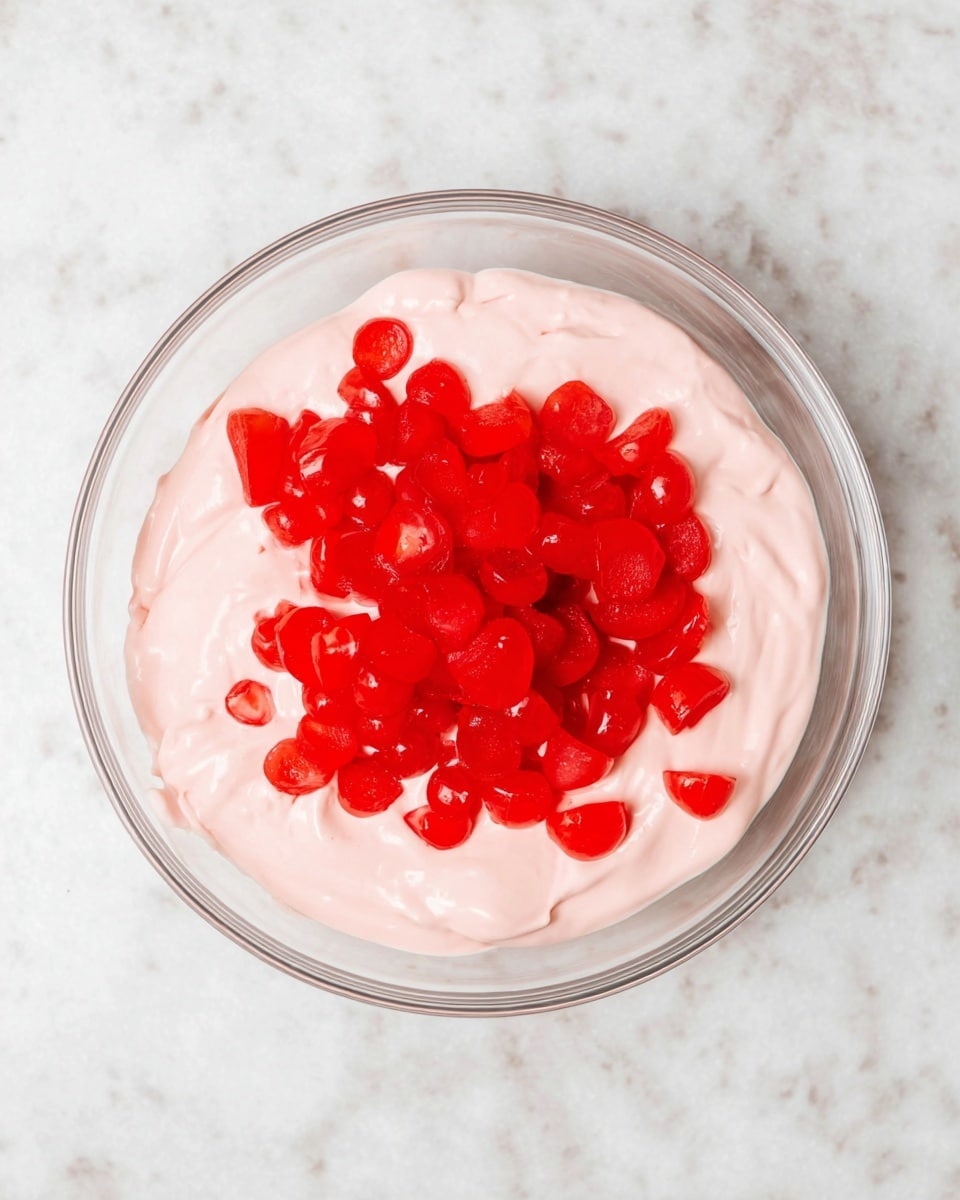 A clear glass bowl is placed on a white marbled surface, filled with a thick, creamy pink mixture that has a smooth texture. On top of the pink layer, there is a generous pile of bright red, small, round, sliced cherries, scattered mainly in the center but some extending toward the edges. The contrast between the light pink cream and vivid red cherries is sharp and colorful. photo taken with an iphone --ar 4:5 --v 7