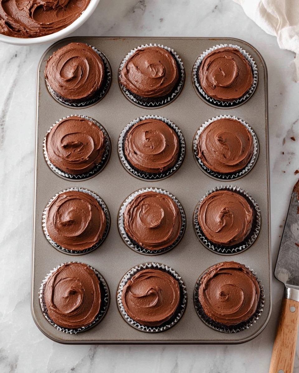 A metal cupcake tray holds twelve chocolate cupcakes, each topped with a smooth layer of dark brown chocolate frosting spread evenly with soft swirls. The cupcakes are dark and moist, visible just under the frosting edges inside white cupcake liners. The tray is placed on a white marbled surface, with part of a bowl filled with chocolate frosting and a knife with frosting resting near the bottom right corner of the image. The overall look is neat, showing the uniform, thick frosting texture on top of every cupcake. photo taken with an iphone --ar 4:5 --v 7