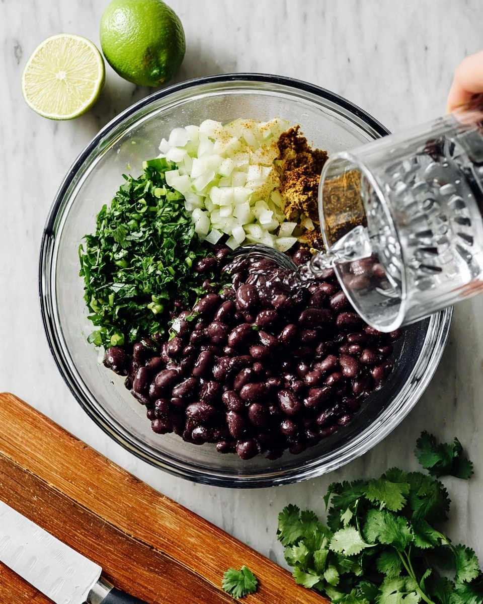 A clear mixing bowl is shown from above with three main layers inside: cooked black beans on the right that are dark purple and shiny, chopped green cilantro leaves on the bottom left, and cooked diced onions mixed with green peppers and spices on the top left. A woman's hand is pouring clear water from a glass onto the black beans. Above the bowl, there is a lime half resting on a clear juicer with juice marks. Below the bowl, on a wooden board, are loose cilantro leaves and a white knife with a brown handle. The surface is a white marbled texture. Photo taken with an iphone --ar 4:5 --v 7