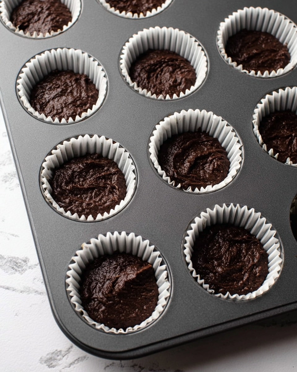A dark brown chocolate batter is placed inside white paper cupcake liners that are set in a dark gray muffin tray. The batter looks thick and slightly uneven in texture, filling each liner about halfway. The tray and liners are arranged neatly, and it is all placed on a white marbled surface. Photo taken with an iphone --ar 4:5 --v 7