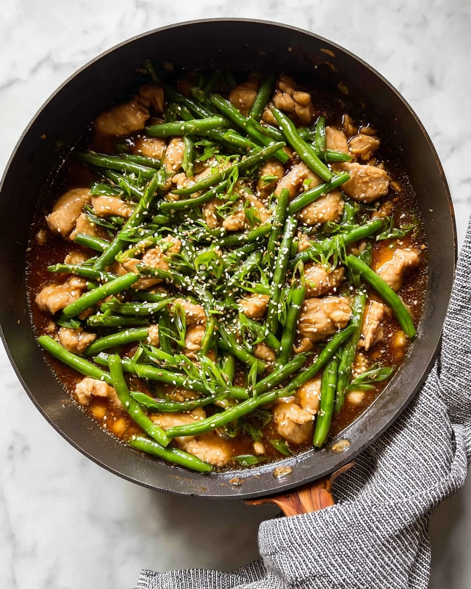 A close-up top view of a dark pan filled with a stir fry dish showing three main layers: the bottom layer has a shiny brown sauce that coats the ingredients, the middle layer is made of bright green green beans that look firm and fresh, spread evenly across the pan, and the top layer includes light brown pieces of cooked chicken along with chopped green onions sprinkled all over and a few sesame seeds scattered on top. The handle of the pan is wrapped in a gray and white striped kitchen towel, and the pan sits on a white marbled surface. photo taken with an iphone --ar 4:5 --v 7