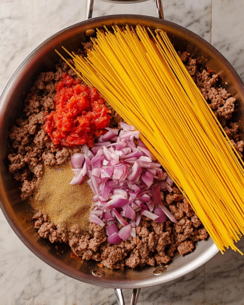 A silver pan filled with a mixture of brown cooked ground meat with pieces of purple onion spread evenly on the bottom layer. On top, there is a bright yellow bundle of uncooked spaghetti arranged on the right side. On the left side, a small pile of red chopped tomatoes sits next to a heap of light brown spice powder. The pan rests on a white marbled surface. photo taken with an iphone --ar 4:5 --v 7