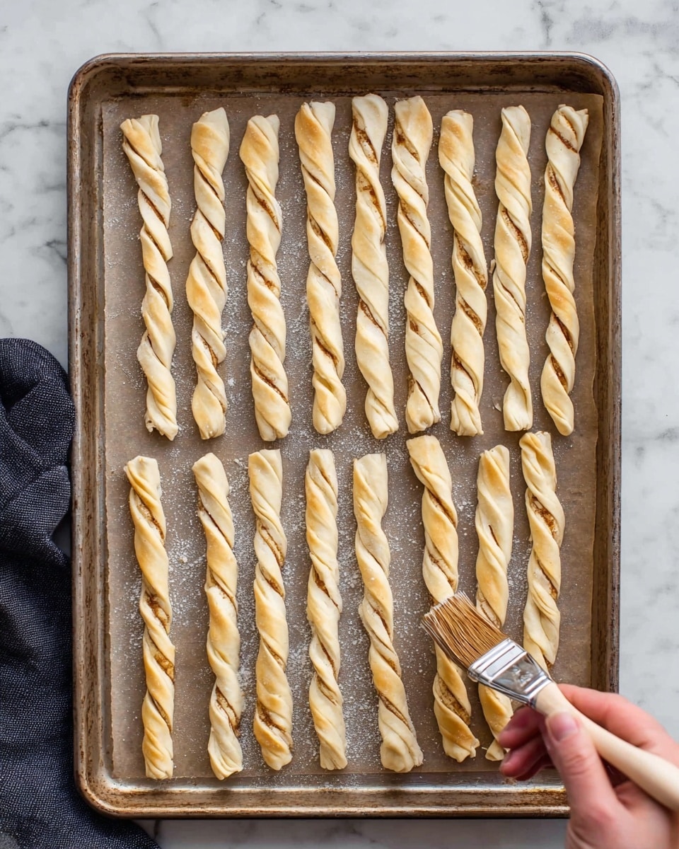 On a baking tray covered with parchment paper, there are twenty twisted dough sticks arranged in four neat rows, each stick having a light golden color with visible cinnamon swirls inside the twists. The dough looks smooth and slightly shiny, likely due to a brush of egg wash being applied by a woman's hand holding a pastry brush at the bottom right corner. The tray rests on a white marbled surface, and a dark cloth is partially visible on the left side. The overall scene is well-lit, showing fine dough texture and subtle browning spots on the baking sheet photo taken with an iphone --ar 4:5 --v 7