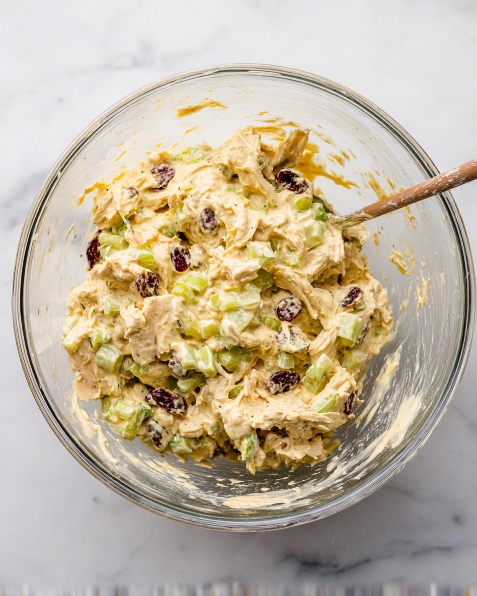 In the clear glass mixing bowl, there is a creamy mixture with visible layers of light yellow dressing coating small pieces of light tan shredded chicken, bright green celery chunks, and dark red dried cranberries. The texture looks thick and rich, with the ingredients evenly mixed throughout. The bowl sits on a white marbled surface, and no woman's hand is visible. photo taken with an iphone --ar 4:5 --v 7