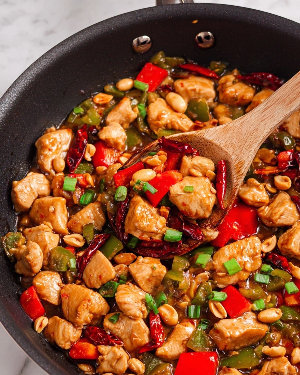 This image shows a close-up of a black skillet filled with a stir-fry dish. There are many chunky pieces of cooked chicken in light brown sauce mixed with bright red and green bell pepper pieces, whole dried red chili peppers, and light brown peanuts. The sauce looks shiny and coats all the ingredients giving a slightly wet texture. Chopped green onions are sprinkled on top adding fresh green color. A wooden spoon scoops a portion of the dish from the skillet, holding several chicken chunks, peppers, peanuts, and green onion pieces. The skillet rests on a white marbled surface. photo taken with an iphone --ar 4:5 --v 7