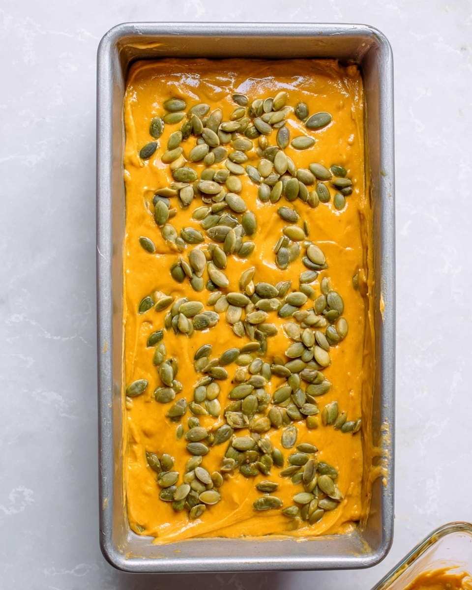 A rectangular silver baking pan filled with a thick, smooth layer of bright orange batter, evenly spread across the pan. Scattered on top is a generous amount of green pumpkin seeds, adding texture and color contrast over the bright orange surface. The pan rests on a white marbled textured surface, and part of a clear container with the same orange batter is visible at the bottom right corner. Photo taken with an iphone --ar 4:5 --v 7