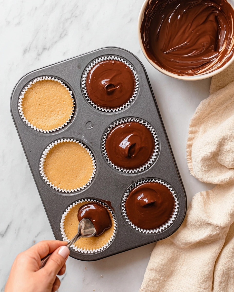 A dark grey muffin pan holds six cupcakes in white paper liners on a white marbled surface. Three cupcakes are plain with a light yellow, slightly cracked top, while the other three are covered with a thick, smooth chocolate layer that shines under the light. A woman's hand is holding a spoon, spreading chocolate evenly on one of the plain cupcakes. Nearby, a white bowl filled with shiny chocolate sits partially in the frame. A cream-colored cloth with soft folds lies at the top right corner. photo taken with an iphone --ar 4:5 --v 7