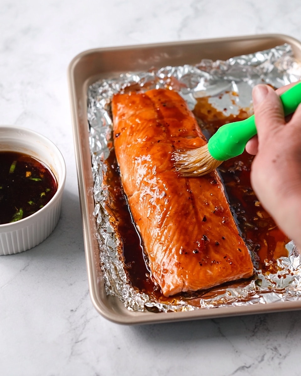 A shiny, orange-brown cooked salmon fillet sits on a tray lined with crinkled silver foil. The fillet has a smooth, glossy texture from the sauce brushed on top, showing some darker caramelized spots. A woman's hand is brushing the surface with a green brush, adding more sauce. To the left side, there is a white bowl filled with dark brown sauce. All of this is set on a white marbled surface. photo taken with an iphone --ar 4:5 --v 7