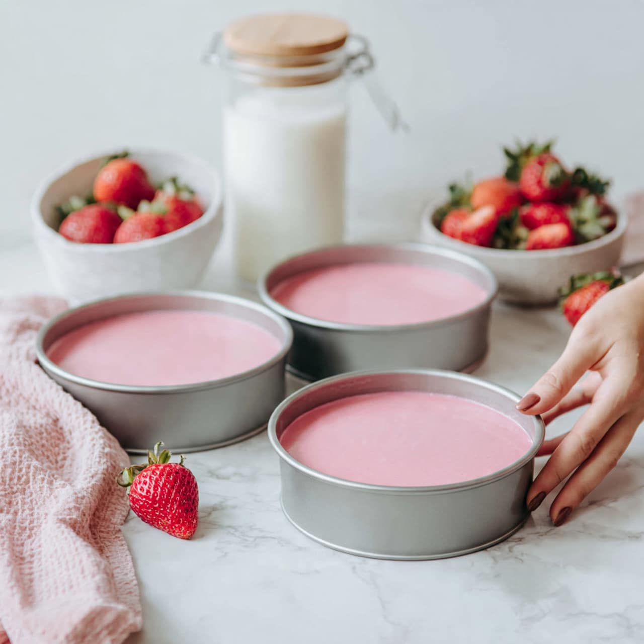 The image shows three round metal bowls filled with smooth, light pink batter spread evenly inside each bowl. Next to them are a white bowl filled with fresh red strawberries, a glass jar with a wooden lid, and a clear glass of milk. A woman's hand is holding a fresh strawberry above a small white bowl filled with more strawberries. All items are placed on a white marbled surface, creating a fresh and clean look. photo taken with an iphone --ar 4:5 --v 7