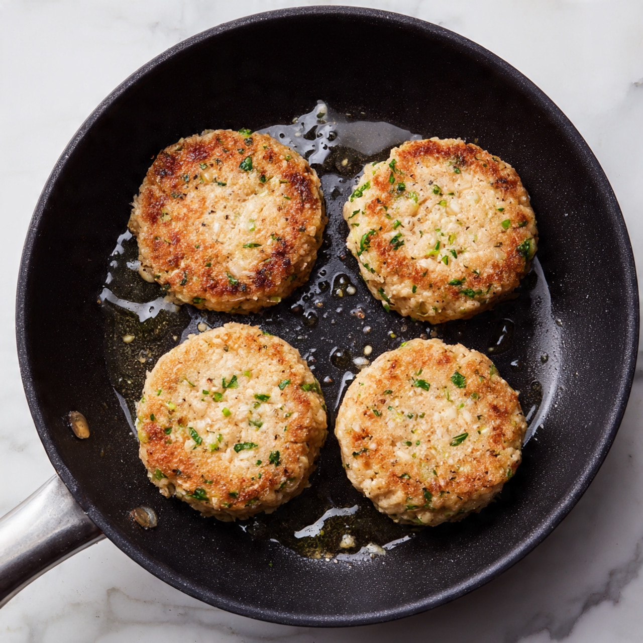 A close-up top view shows four round, pale beige patties with small green herb pieces mixed in, frying in a black pan with a few drops of oil. The patties have a slightly rough texture and are spaced evenly in the pan, which sits on a white marbled surface photo taken with an iphone --ar 4:5 --v 7