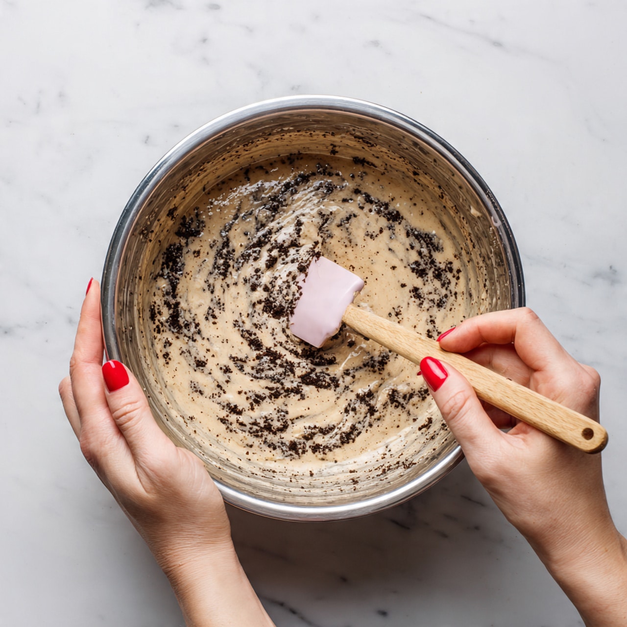 A close-up top view of a shiny silver mixing bowl held by a woman's hand with red nail polish on a white marbled surface, filled with light beige creamy batter mixed with dark brown crushed cookie pieces scattered evenly inside. A woman's hand with red nail polish holds a light pink spatula with a wooden handle, stirring the mixture in the bowl. photo taken with an iphone --ar 4:5 --v 7