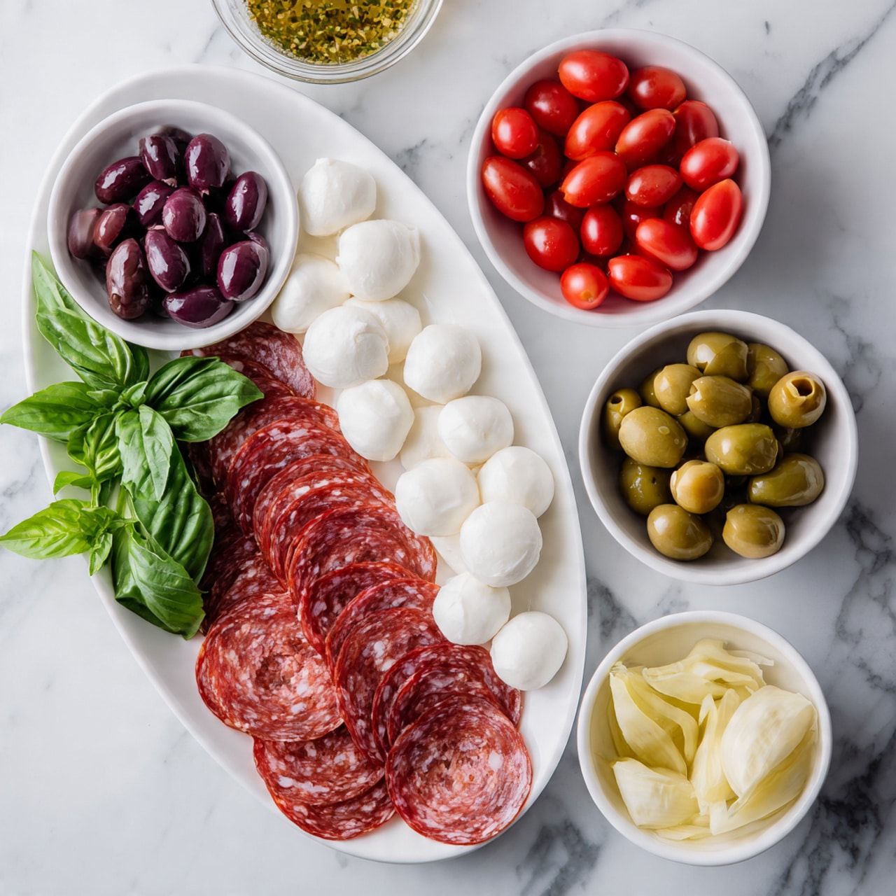 The image shows several small white bowls and one white oval plate arranged on a white marbled surface. The white oval plate at the bottom left is filled with thin, round slices of pink and white speckled salami, layered neatly. To the top left, there is a small white bowl with dark purple kalamata olives. In the center top, a larger white bowl holds bright red, shiny grape tomatoes. To the right of the tomatoes, a white bowl is full of small, smooth white mozzarella balls. Below the mozzarella, a small round clear glass bowl contains a golden-yellow Italian dressing with visible herbs. Near the bottom right, a white bowl is filled with light yellow chopped artichokes, and next to it, another white bowl holds medium green Castelvetrano olives. Fresh dark green basil leaves with prominent veins lay in the middle left, partially overlapping the salami plate. photo taken with an iphone --ar 4:5 --v 7