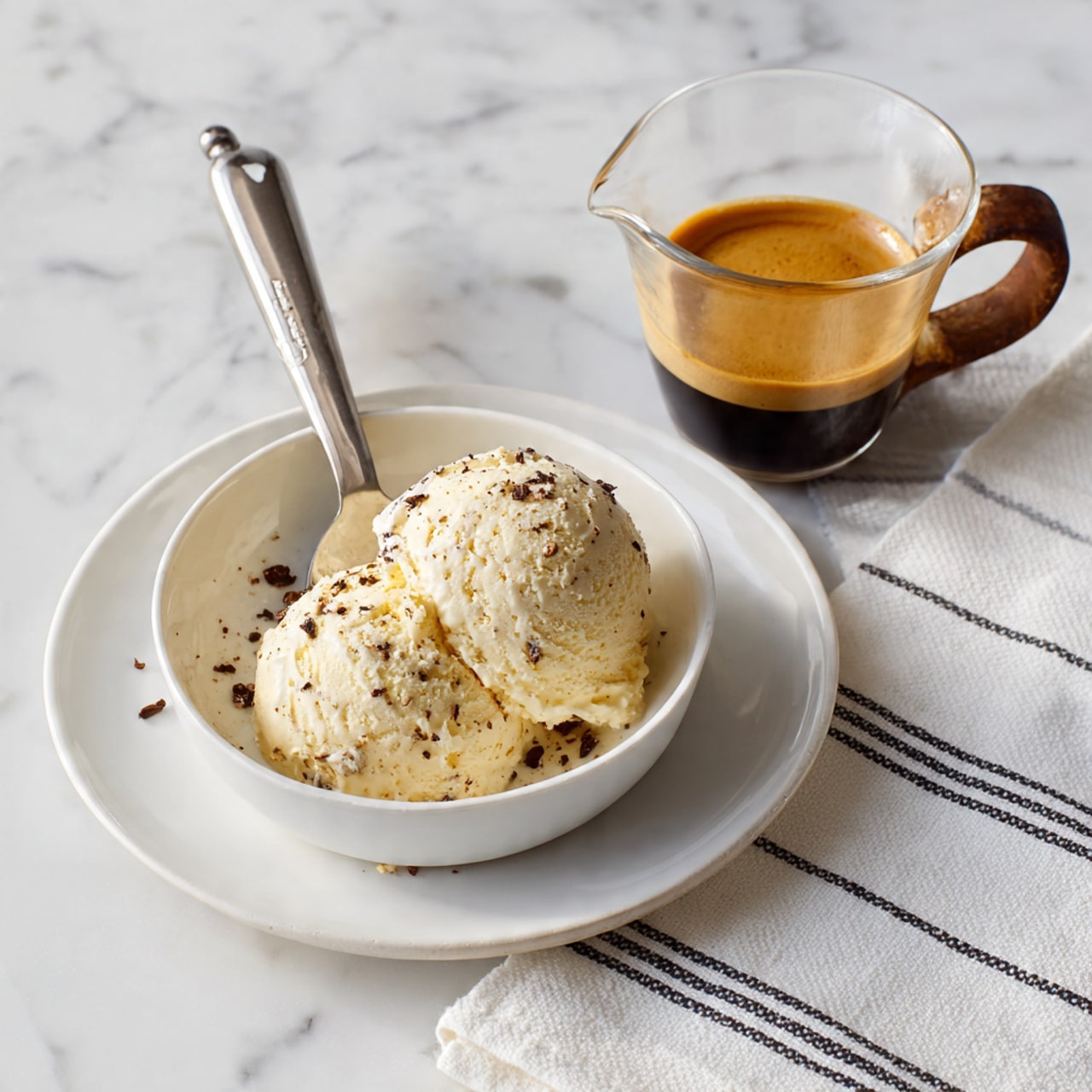 The image shows a white plate with a scoop of vanilla ice cream mixed with small dark bits, held by a metal ice cream scooper resting on the plate. Above the plate, a small glass jug with a dark wooden handle contains golden-brown espresso. The scene is set on a white marbled surface with a white cloth featuring thin black stripes on the right side. Photo taken with an iphone --ar 4:5 --v 7