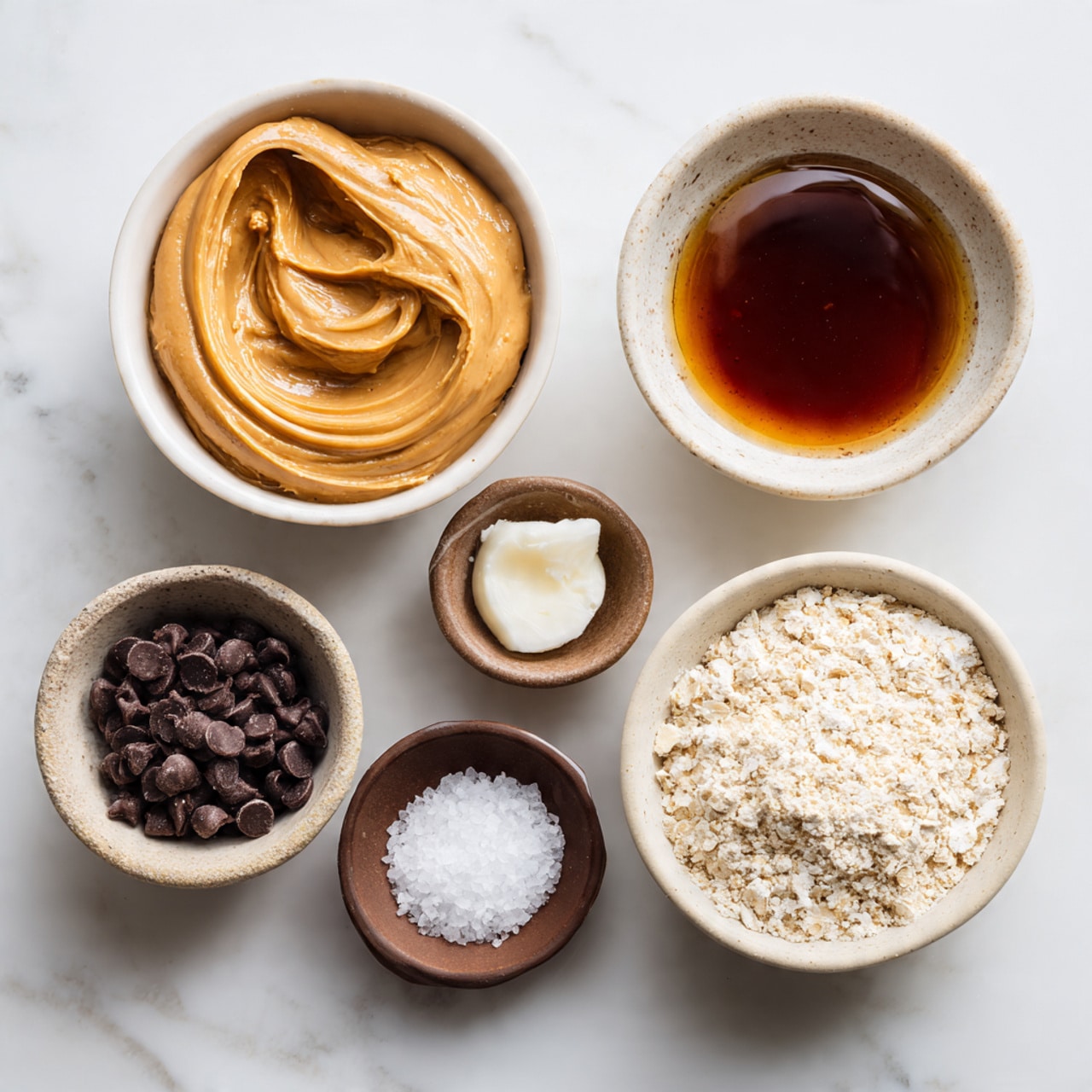 Five small white bowls sit on a white marbled surface, each holding a different ingredient. The largest bowl at the top left is filled with smooth, light brown natural peanut butter with a creamy texture and slight swirls. To its right, a smaller bowl holds dark amber maple syrup with a glossy and translucent surface. Below the peanut butter, another small bowl contains dark brown chocolate chips with a small dollop of white coconut oil on top, showing a contrast between the solid chips and soft oil. To the right of that is a slightly bigger bowl filled with pale beige oat flour, finely ground with a soft texture. At the bottom center, a tiny brown bowl holds coarse white salt crystals. photo taken with an iphone --ar 4:5 --v 7