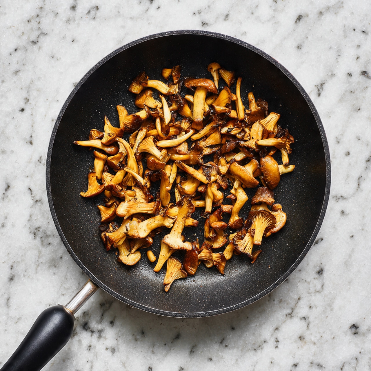 The image shows a non-stick black frying pan with small golden-brown cooked mushrooms scattered inside. The mushrooms are of different shapes and sizes, some long and thin while others are short and wider, with a lightly toasted texture on their surfaces. The pan sits on a white marbled surface with a speckled finish, and the pan’s handle is black, extending out of the top of the frame. photo taken with an iphone --ar 4:5 --v 7