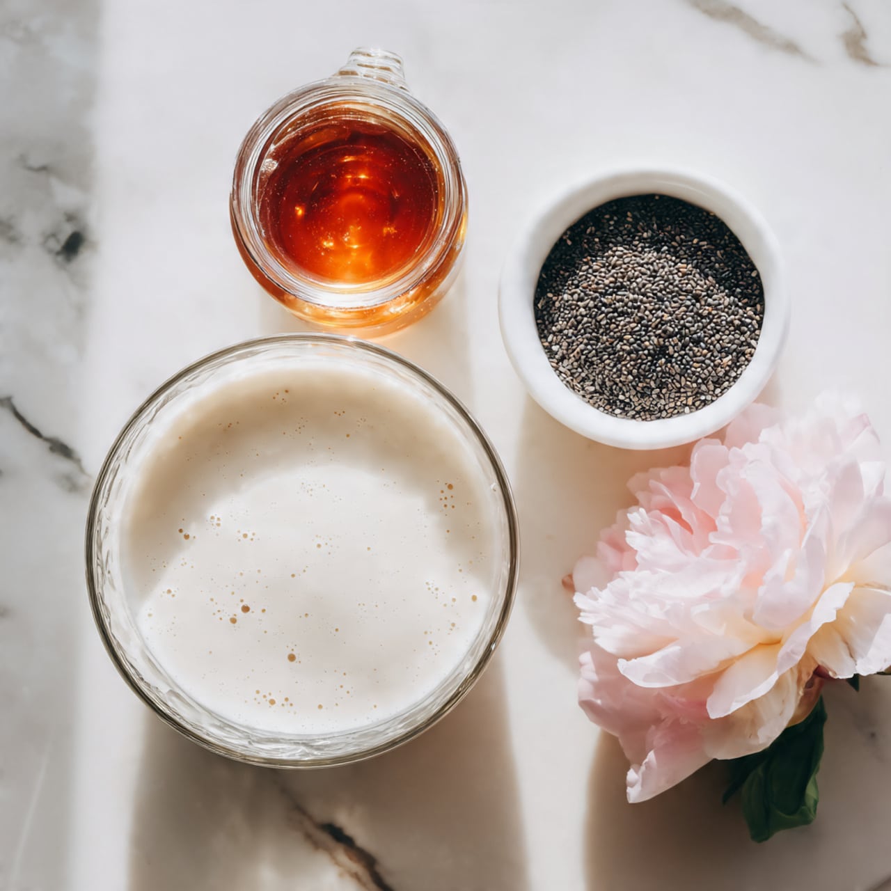 The image shows two clear glass jars filled with layered chia pudding on a white marbled board. The pudding has two layers: a bottom layer of creamy white chia seeds soaked in milk, dotted with tiny black seeds, and a thick top layer of bright red strawberry puree with sliced strawberries visible inside and on top. The jars have wide openings and rounded sides. A fresh whole strawberry with green leaves and a soft pink rose petal are placed nearby on the white marbled surface. The background is a soft pink color. Photo taken with an iphone --ar 4:5 --v 7