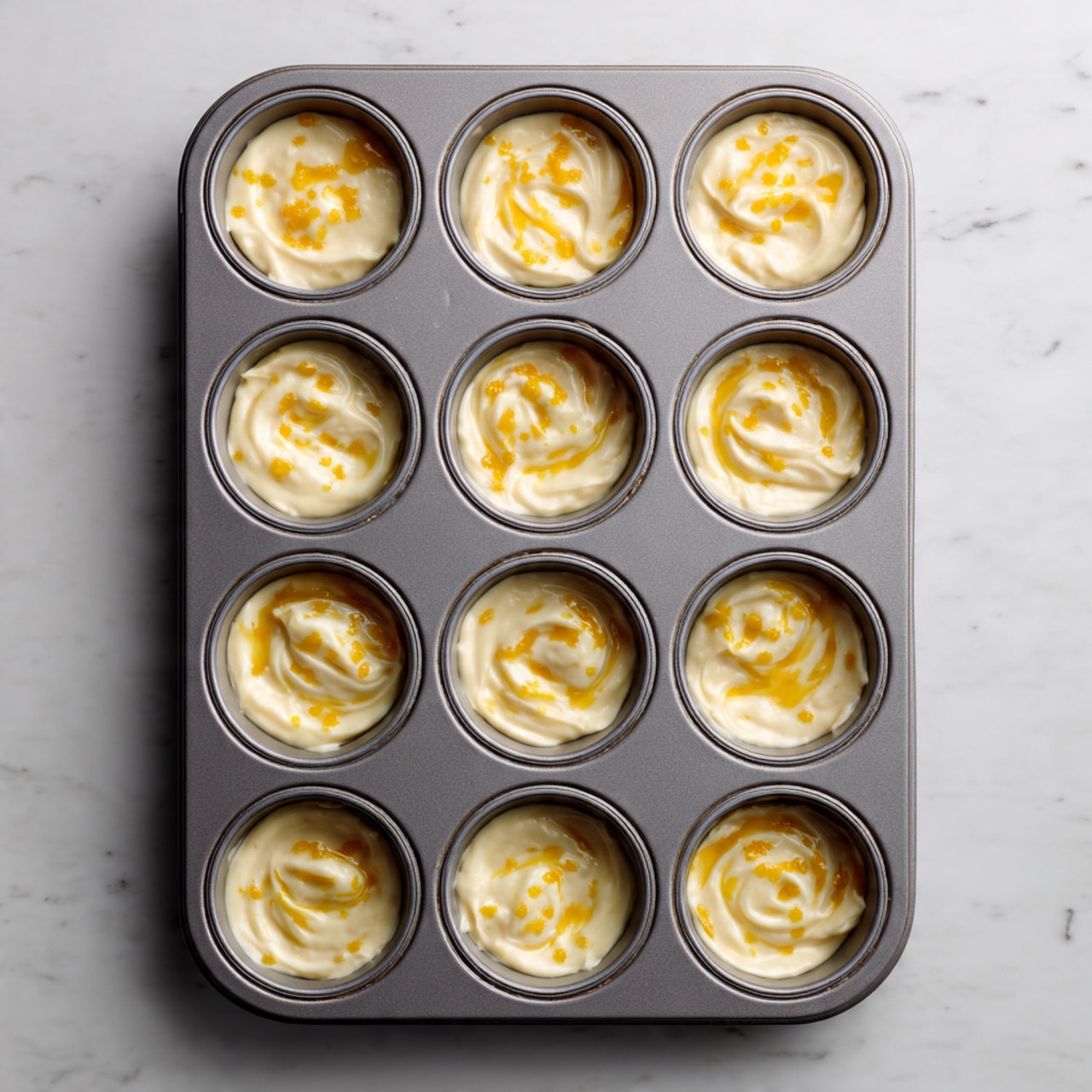 A gray metal muffin tray holds twelve unbaked dough cups, each filled with a smooth, light beige batter topped with irregular golden-yellow drizzles that spread unevenly over the surface. The batter in each cup looks soft with gentle folds and slight indentations, showing a creamy texture. The tray sits on a white marbled surface, and the image is taken from above, showing the uniform arrangement of the cups in three rows of four. photo taken with an iphone --ar 4:5 --v 7