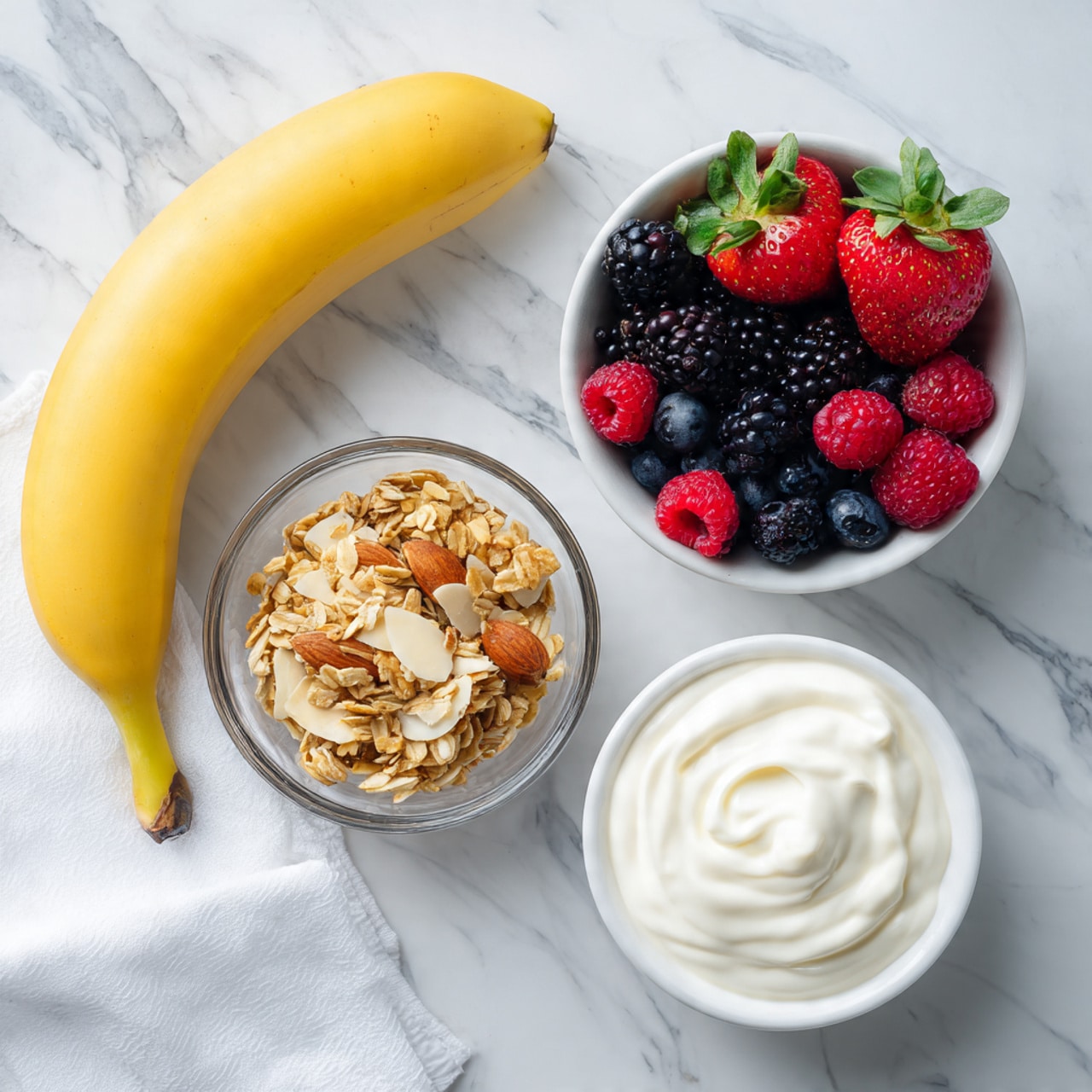 A bright yellow banana lies diagonally on the top left of the image on a white marbled surface. To the right of the banana, there is a white bowl filled with fresh berries that include red strawberries with green leaves, dark blackberries, small blue blueberries, and red raspberries. Below the berry bowl, there is a smaller clear glass bowl filled with golden-brown granola mixed with sliced almonds. To the right of the granola bowl, a white bowl contains smooth, creamy Greek yogurt with gentle swirls on the surface. All items are arranged neatly on the white marbled background with a white cloth visible at the bottom left corner of the image. Photo taken with an iphone --ar 4:5 --v 7