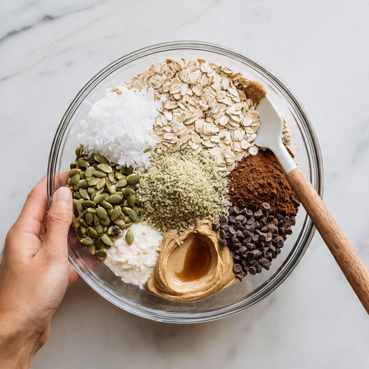 A clear glass bowl contains eight distinct layers of ingredients gathered for mixing on a white marbled surface. Starting from the top left, there is a dollop of white coconut oil with a smooth texture, next to a pile of light beige rolled oats with a rough texture, and beside that a light greenish-beige cluster of hemp seeds. To the right of the hemp seeds is a mound of dark brown cocoa powder with a fine texture. Below the cocoa powder is smooth peanut butter with a creamy light brown color, next to which is a small pool of dark brown liquid vanilla extract. At the bottom left is a dollop of pale beige mashed banana, topped partially by a layer of green pumpkin seeds with a shiny texture. In the center rests a pile of small, dark brown chocolate chips. A woman's hand is holding a spatula with a white silicone head and wooden handle resting on the edge of the bowl. Photo taken with an iphone --ar 4:5 --v 7