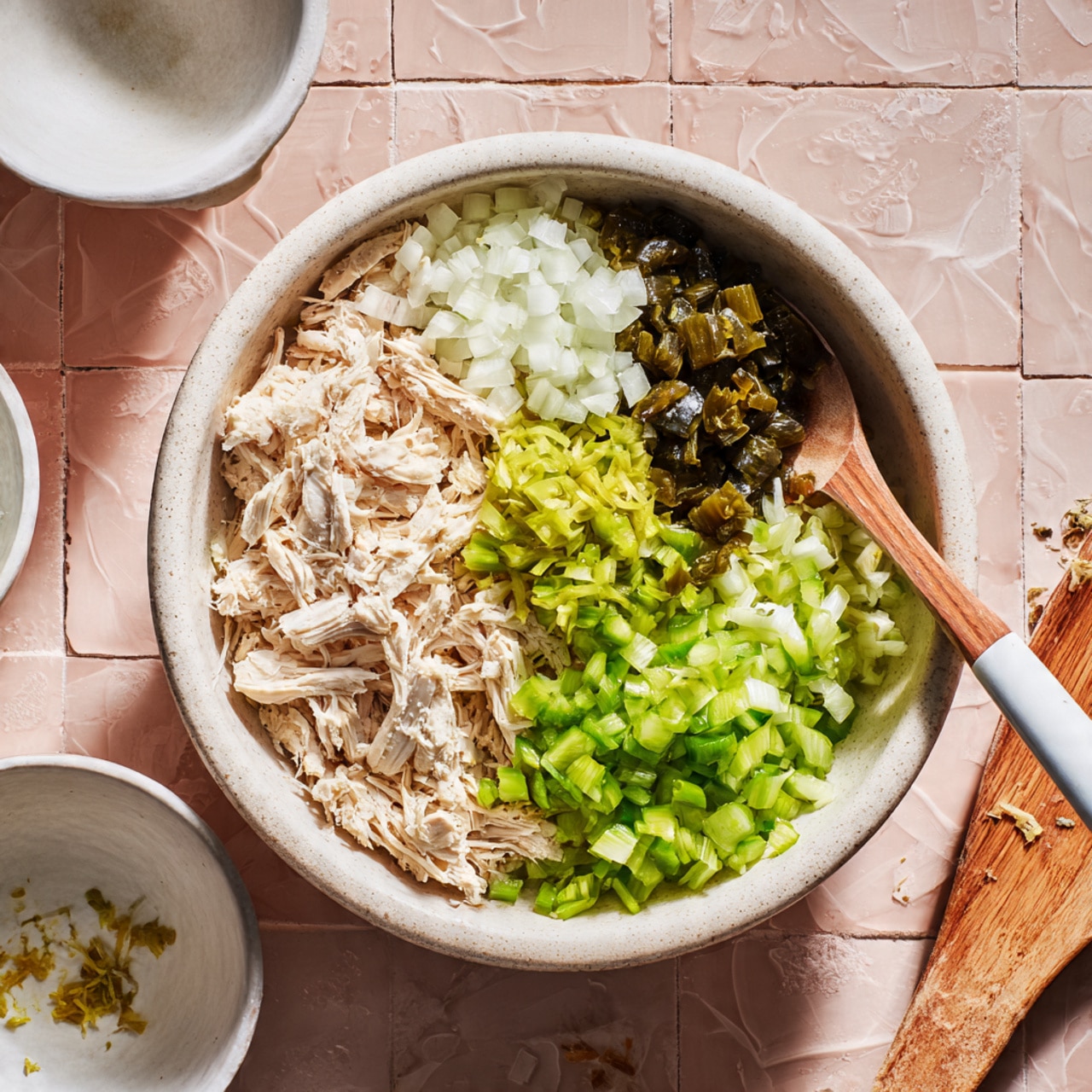 In a white bowl with a lined texture, there are four main layers starting with a base of shredded pale cooked chicken. On the left side, finely chopped white onion adds a light texture. Above the chicken is a small pile of dark green diced pickles, and to the right, a heap of chopped bright green celery. On top of the chicken in the center is a small pile of finely chopped green peppers. A wooden spoon with a white handle rests partially in the bowl on the left side. The bowl is placed on a soft pink tiled surface with a textured pattern. Nearby, there are small white bowls with some remnants of ingredients and a wooden spatula at the bottom right. photo taken with an iphone --ar 4:5 --v 7