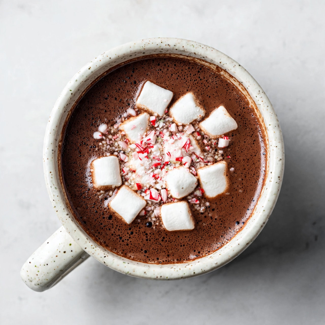 A white speckled mug filled with dark brown hot chocolate, topped with a layer of small white marshmallows and sprinkled with crushed red and white peppermint pieces in the center, sits on a matching white speckled saucer placed on a white marbled surface. Nearby, a silver spoon lies to the left and some crushed peppermint bits scatter around, with a soft gray cloth partially visible to the right. A second mug with a similar drink and marshmallows is partially seen at the top left corner. Photo taken with an iphone --ar 4:5 --v 7