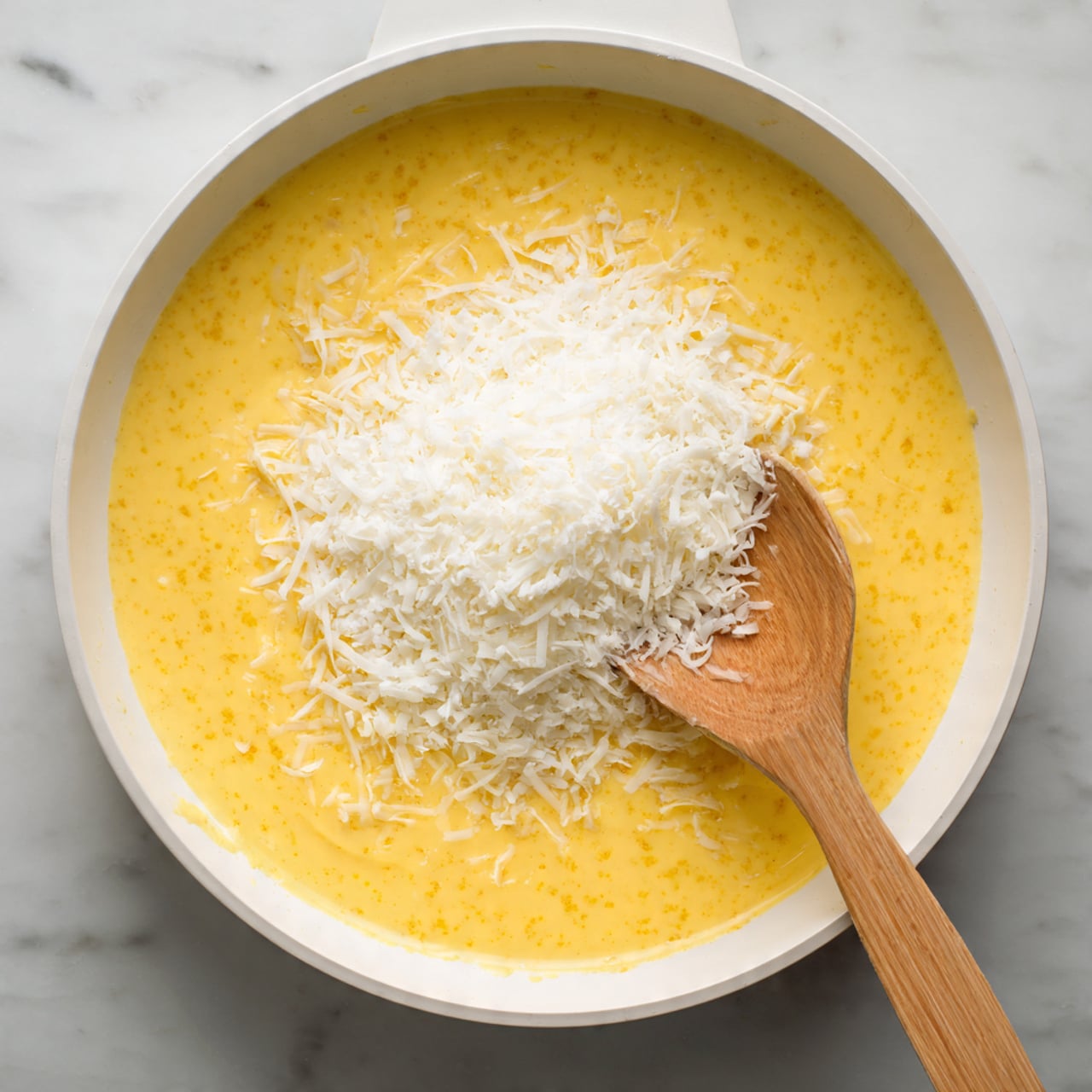 A white round shallow pan filled with a bright yellow creamy sauce that has small textured bits throughout. On top of the sauce sits a pile of finely grated white cheese. A wooden spoon rests on the right side of the pan, partially dipped in the sauce with some cheese on it. The pan is placed on a white marbled surface. Photo taken with an iphone --ar 4:5 --v 7