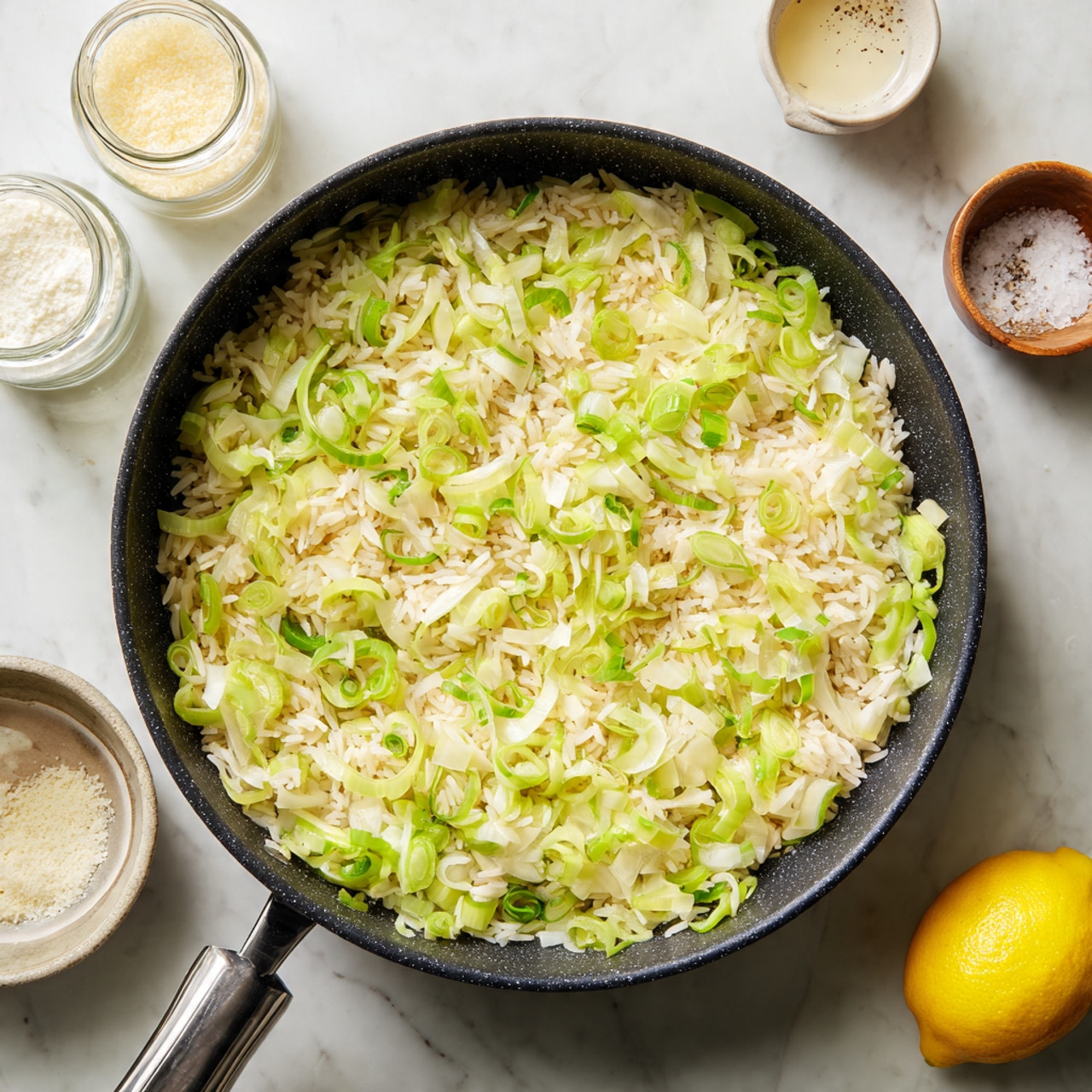 A large black speckled pan sits on a white marbled surface, filled with one layer of uncooked rice mixed evenly with thinly sliced light green and bright green vegetables, likely leeks or scallions, forming a scattered texture. Above the pan, jars and small bowls with grated cheese, a creamy liquid, and salt with pepper are placed on the white marbled surface, along with half a lemon at the bottom right corner. The colors are mainly white, pale yellow, and green against the black pan and white marble background. photo taken with an iphone --ar 4:5 --v 7