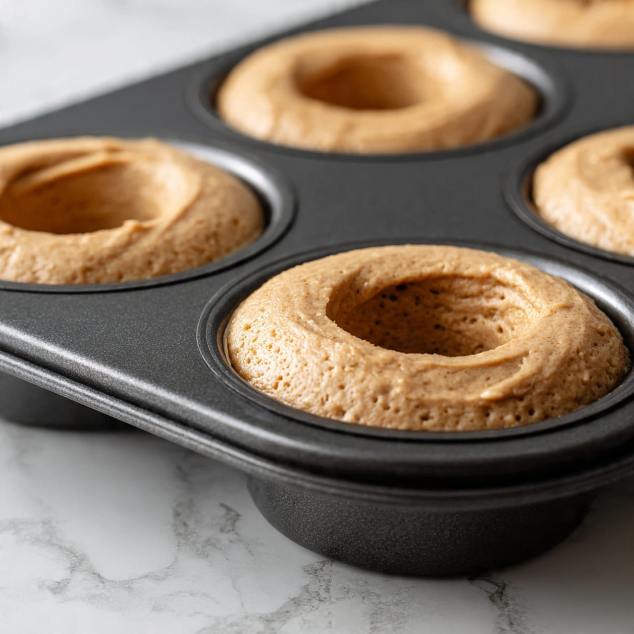 The image shows several small round pastries with a light brown dough base and a glossy caramel-like frosting layer on top. Each pastry has spots of darker brown, adding texture to the shiny glaze. On top of the frosting, there are slices of banana with a light yellow color and some cinnamon powder sprinkled over them. The pastries are placed on a piece of parchment paper on top of a wire rack, with a white marbled surface underneath. One pastry in the background has a bite taken out of it showing a soft and fluffy inside layer. Photo taken with an iphone --ar 4:5 --v 7
