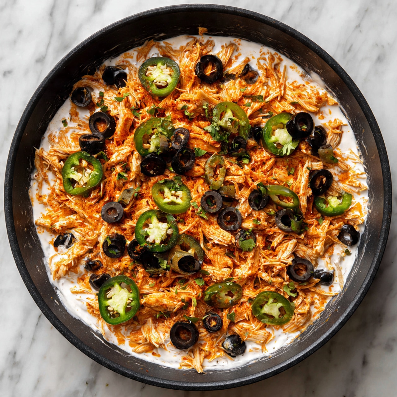 The image shows a black frying pan filled with a layered dish. The bottom layer is white creamy sauce, spread evenly across the pan. On top of this is a layer of shredded orange-colored chicken spread out to cover the sauce. The final layer is scattered with sliced black olives and green jalapeño rings placed evenly all over the chicken. The frying pan is on a surface with a white marbled texture. photo taken with an iphone --ar 4:5 --v 7