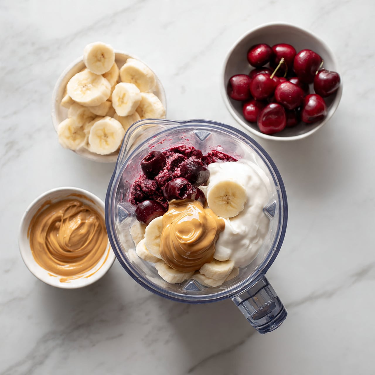 A clear blender jar is shown from above, holding three main layers of ingredients: a base layer of dark red cherries, a middle layer of white yogurt, and a top layer with a peeled banana half placed on the yogurt and a dollop of golden peanut butter sitting beside the banana. Around the blender, a small white bowl with creamy peanut butter and a white bowl with a mix of cherries and sliced banana pieces are arranged, all set on a white marbled surface. photo taken with an iphone --ar 4:5 --v 7