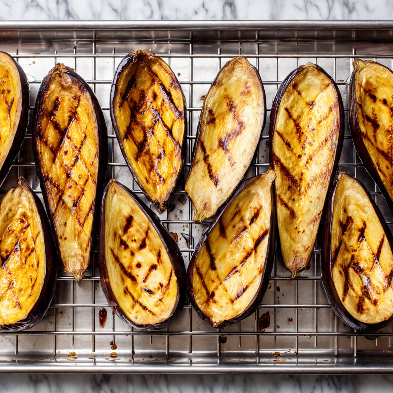 The image shows eight long roasted eggplant halves placed on a shiny metal grill rack. Each eggplant half has golden brown skin with dark brown grilled lines and a few lighter yellow areas where the eggplant flesh is soft. The eggplants have shallow diamond-shaped cuts on their surface, giving texture to the smooth skin. Some oil droplets and small brown charred spots are visible on the metal rack below the eggplants. The background is a white marbled texture. photo taken with an iphone --ar 4:5 --v 7