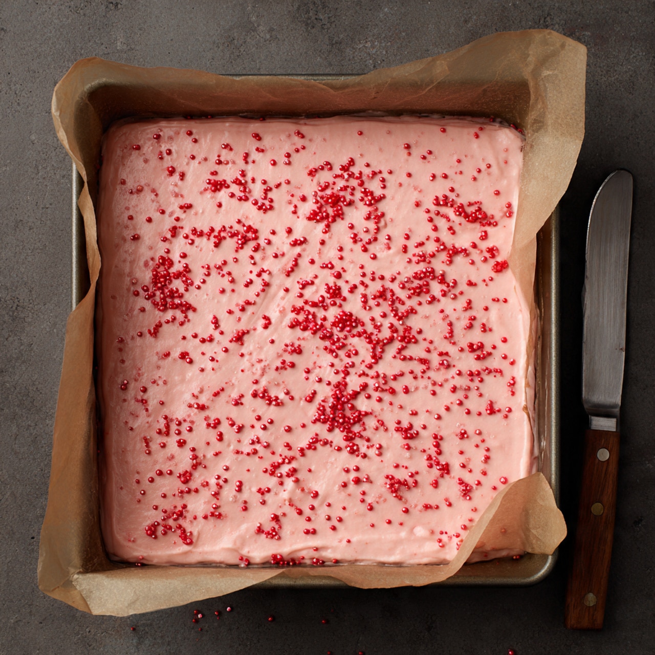A square baking pan lined with light brown parchment paper holds a smooth, pale pink mixture spread evenly to fill the pan. On top, small red sprinkles are scattered all over, creating a textured look with their bright red color against the soft pink surface. The pan sits on a dark countertop with a butter knife nearby. photo taken with an iphone --ar 4:5 --v 7