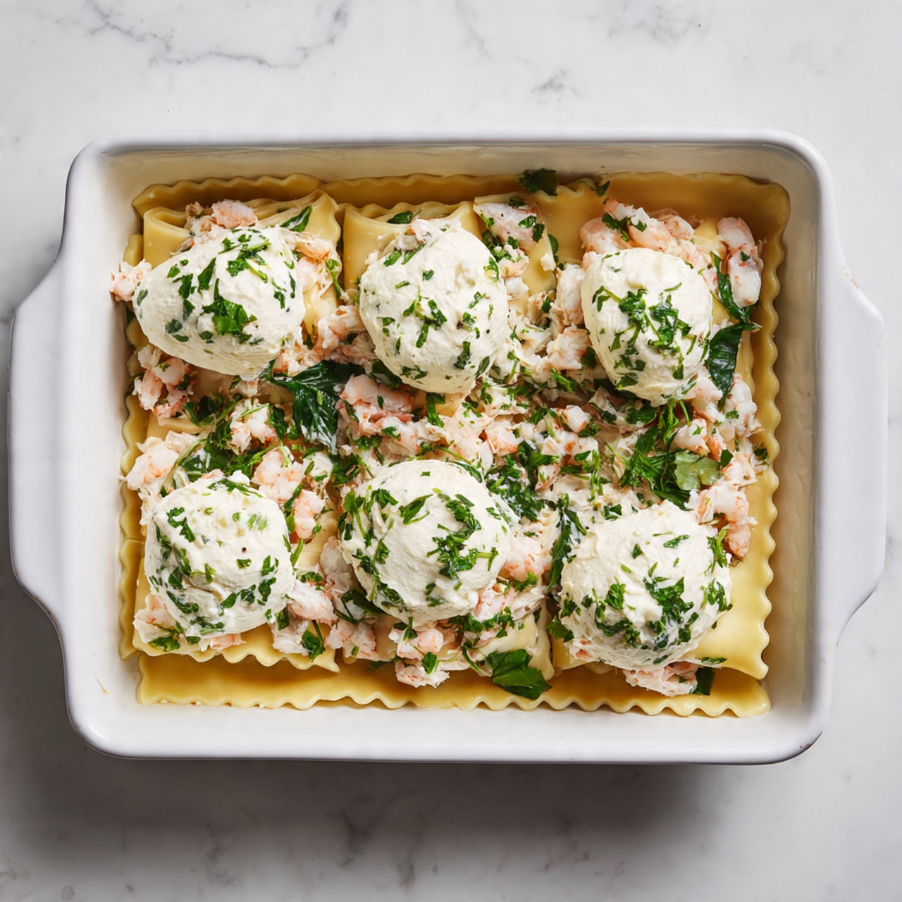 In a white rectangular baking dish, there is one visible layer of uncooked lasagna pasta sheets lining the bottom. On top of the pasta, there is an even layer of small chopped pieces of light pink and white seafood mixed with green spinach leaves. Over this, there are six dollops of white creamy cheese mixed with green herbs placed in a scattered pattern. The dish is set on a white marbled surface. photo taken with an iphone --ar 4:5 --v 7