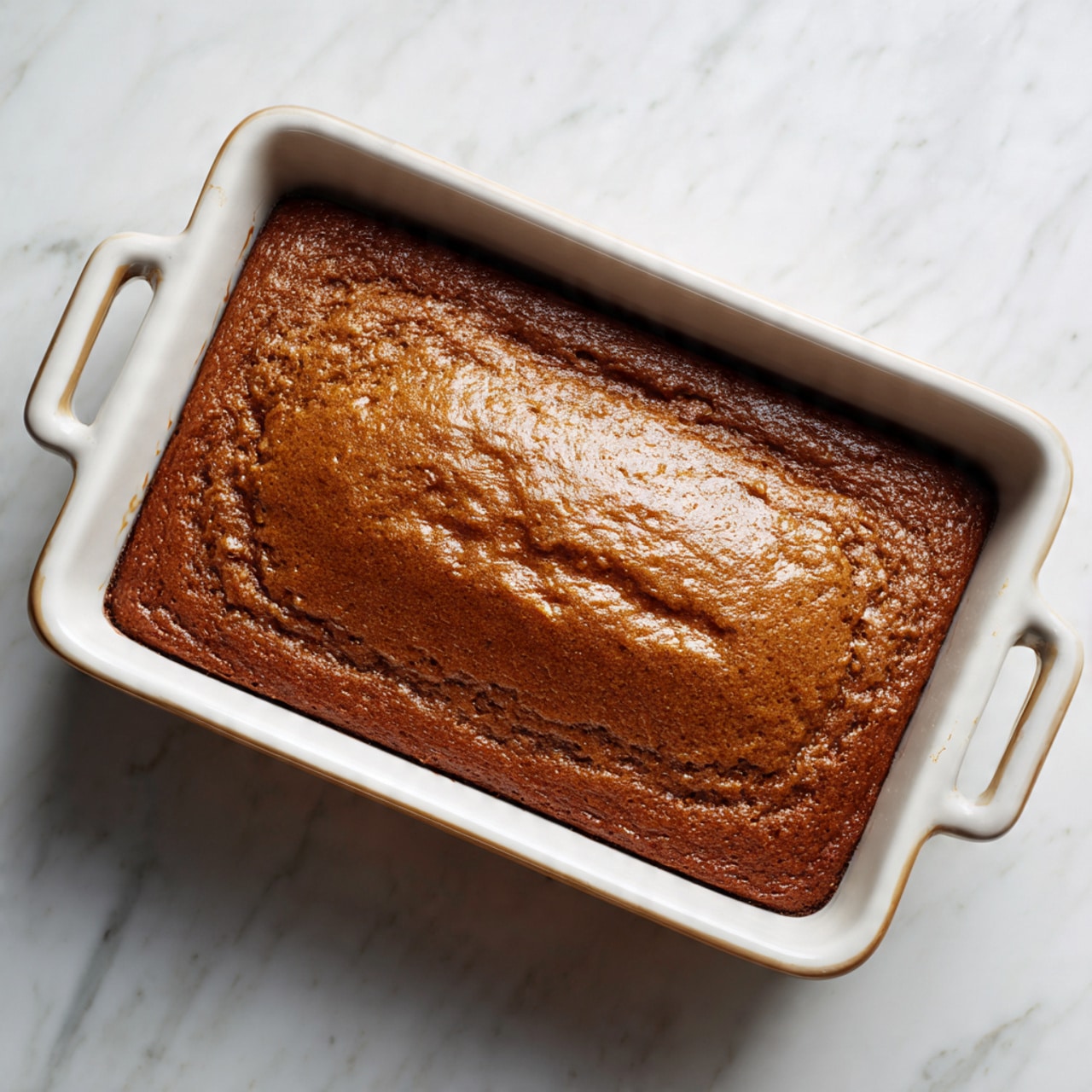 A single large rectangular brown cake with a smooth and slightly textured golden-brown top layer, filling a white ceramic baking dish with handles on each side, placed on a white marbled surface, photo taken with an iphone --ar 4:5 --v 7