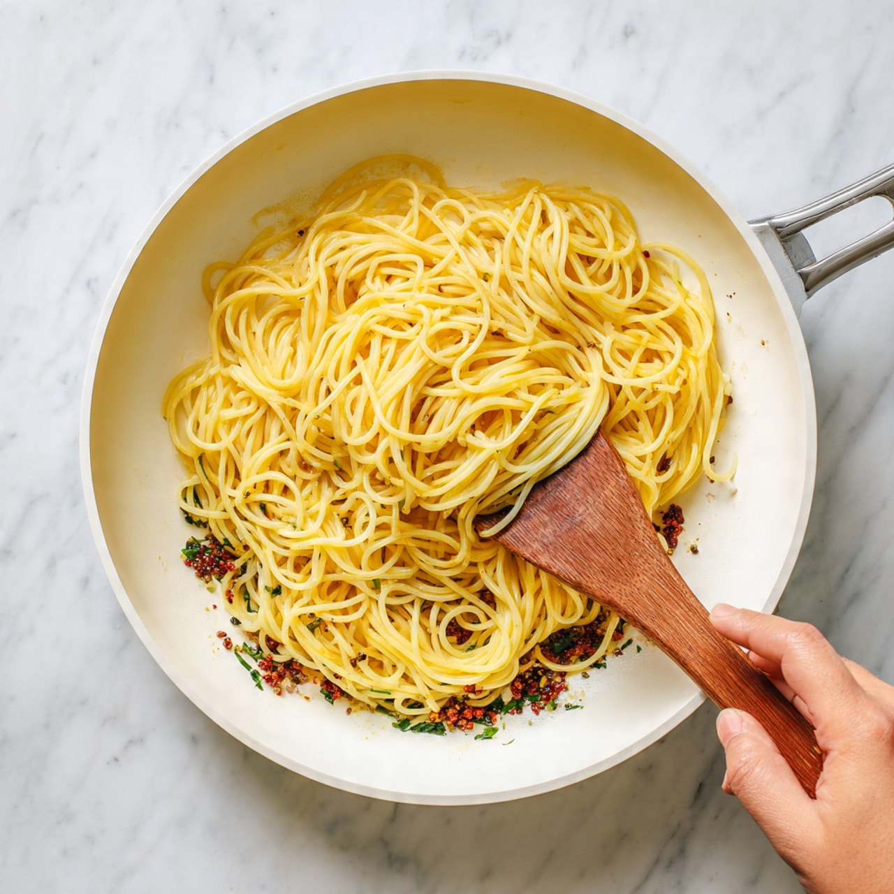 A white pan with a wooden handle holds a single layer of plain yellow spaghetti noodles. Underneath the noodles, there is a thin layer of dark green and brown herbs and spices spread on the bottom of the pan. The pan is set on a white marbled surface. A woman's hand is holding the wooden handle from the top edge of the image photo taken with an iphone --ar 4:5 --v 7