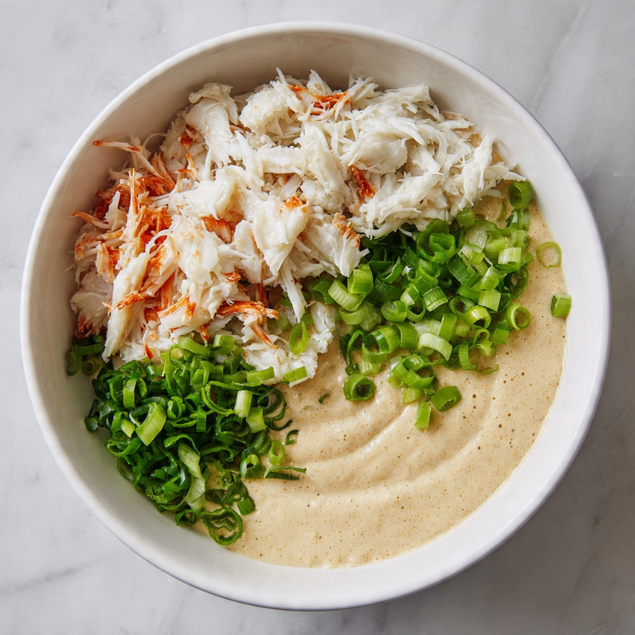 A white bowl sits on a white marbled surface with three separate layers inside. The bottom right side has a thick, creamy light beige sauce with a smooth texture. To the left, there is a pile of white shredded crab meat with some orange pieces mixed in. At the top, fresh bright green chopped scallions add a fresh contrast. The layers are placed side by side without mixing. Photo taken with an iphone --ar 4:5 --v 7