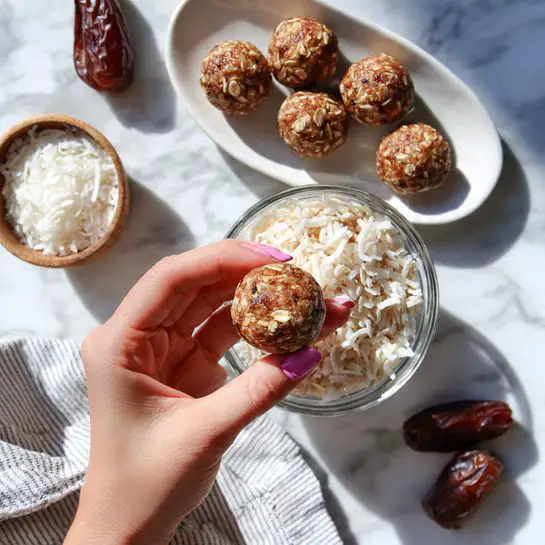 A woman’s hand with pink nail polish holds a small round ball made of brown mixture with visible oats and seeds in front of a clear bowl filled with more of the same mixture. The background is a white marbled surface with some dates scattered around and a white bowl with light brown edges filled with shredded coconut. In the second part of the image, there is a white oval plate with ten brown balls evenly spaced on it, next to the white bowl of shredded coconut, with a small white bowl of dates on the side and a striped cloth near the bottom. The lighting is bright and natural, enhancing the texture and colors of the ingredients photo taken with an iphone --ar 4:5 --v 7