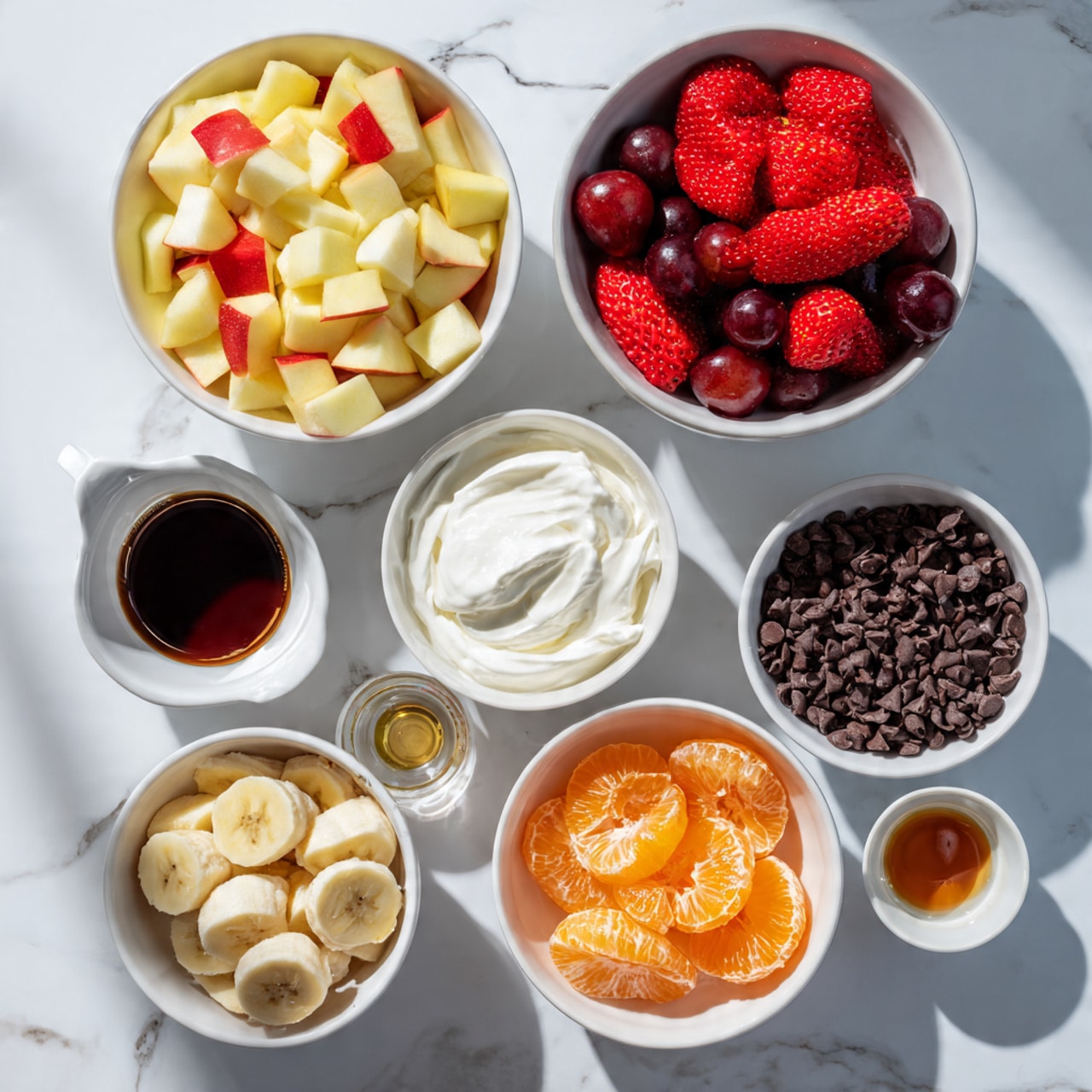The image shows eight white bowls and small containers with different colorful ingredients arranged on a pink tiled surface with visible cracks. Starting from the top right, there is a white bowl filled with bright red halved strawberries. Below it is a smaller bowl filled with dark red halved grapes. To the right of the grapes is a small white container with creamy white heavy whipping cream. Near the center left is a large white bowl full of diced apples with yellow flesh and red skin. To the left of that is a small white jug holding a dark liquid labeled as vanilla extract. Below the apples is a clear small container filled with amber maple syrup. At the bottom left is another white bowl with sliced bananas showing pale yellow flesh and light brown edges. In the bottom center is a white bowl filled with bright orange mandarin orange segments. Lastly, at the bottom right is a small white bowl filled with dark brown chocolate chips. The light and shadows suggest bright natural daylight. Photo taken with an iphone --ar 4:5 --v 7